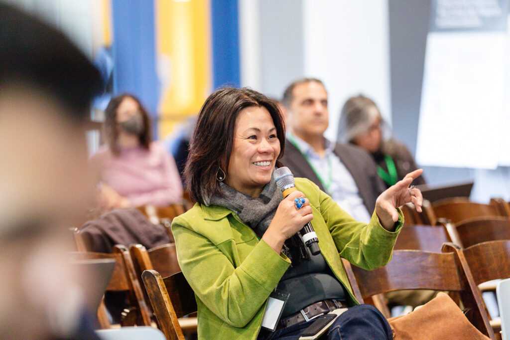 Woman in audience wearing green jacket and holding microphone asks question